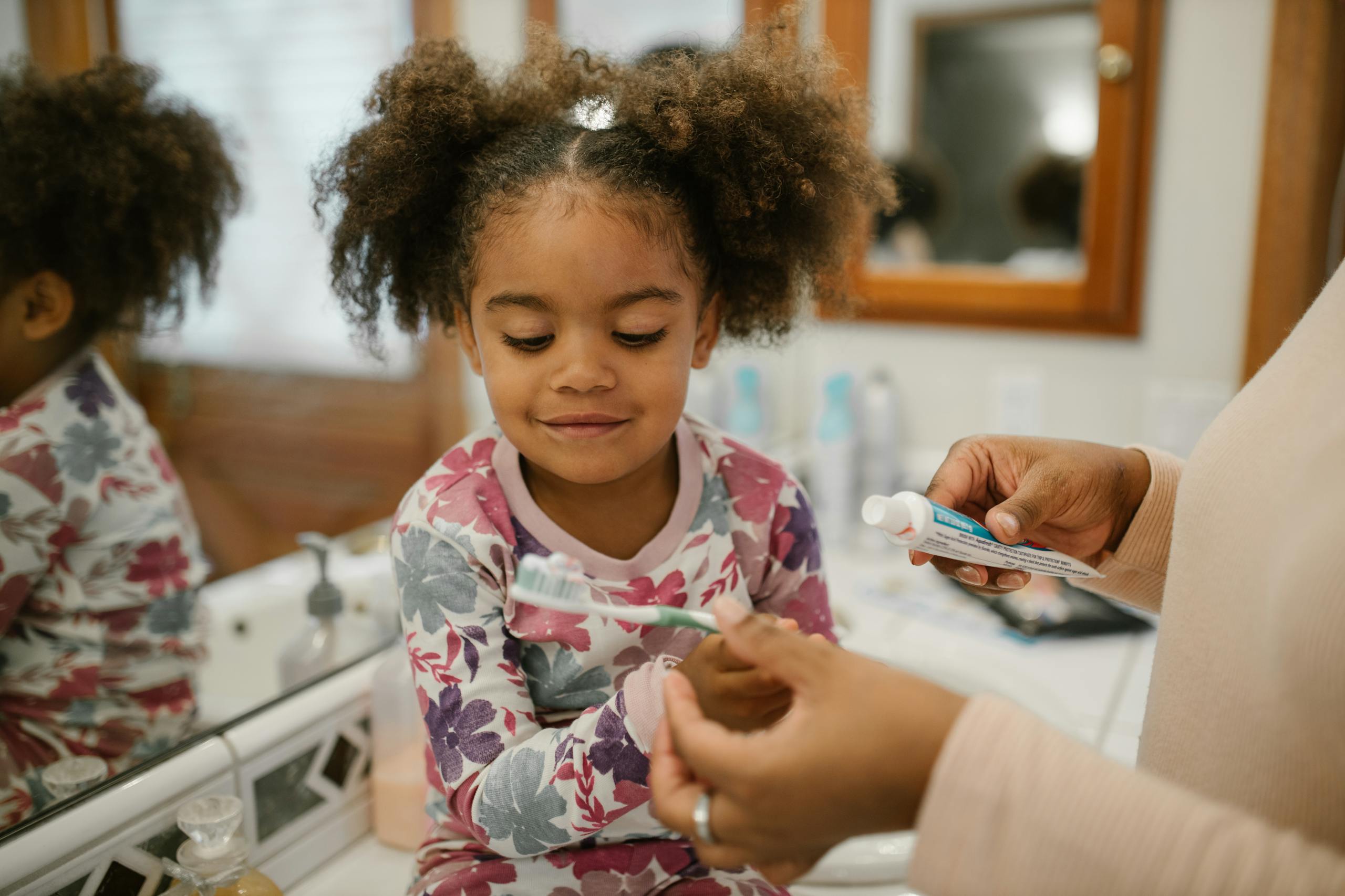 A young girl on a bathroom counter as an adult applies toothpaste to her toothbrush, emphasizing pediatric dental care