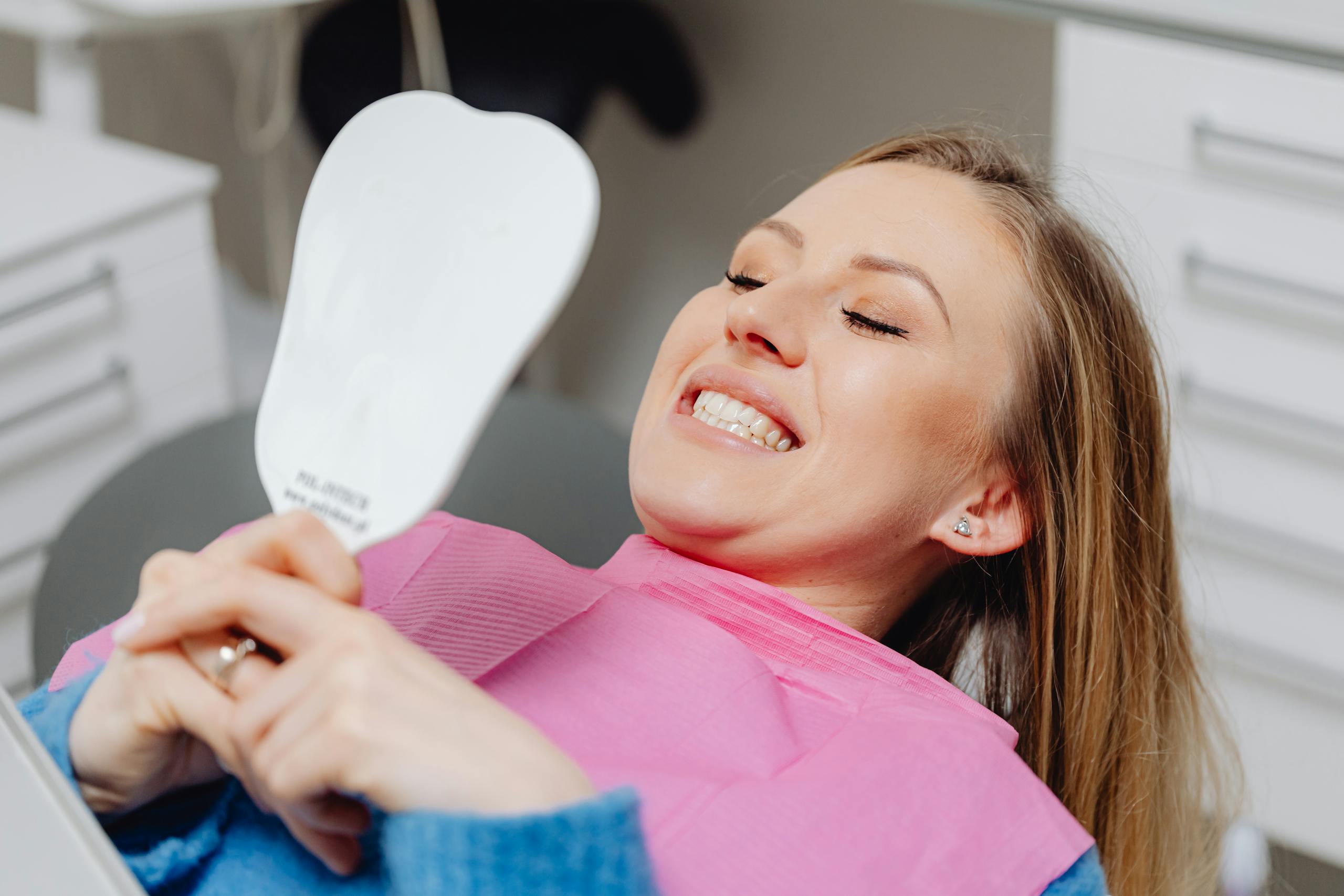 A woman reclines in a dental chair, examining her teeth in a mirror, illustrating satisfaction after a sealant application