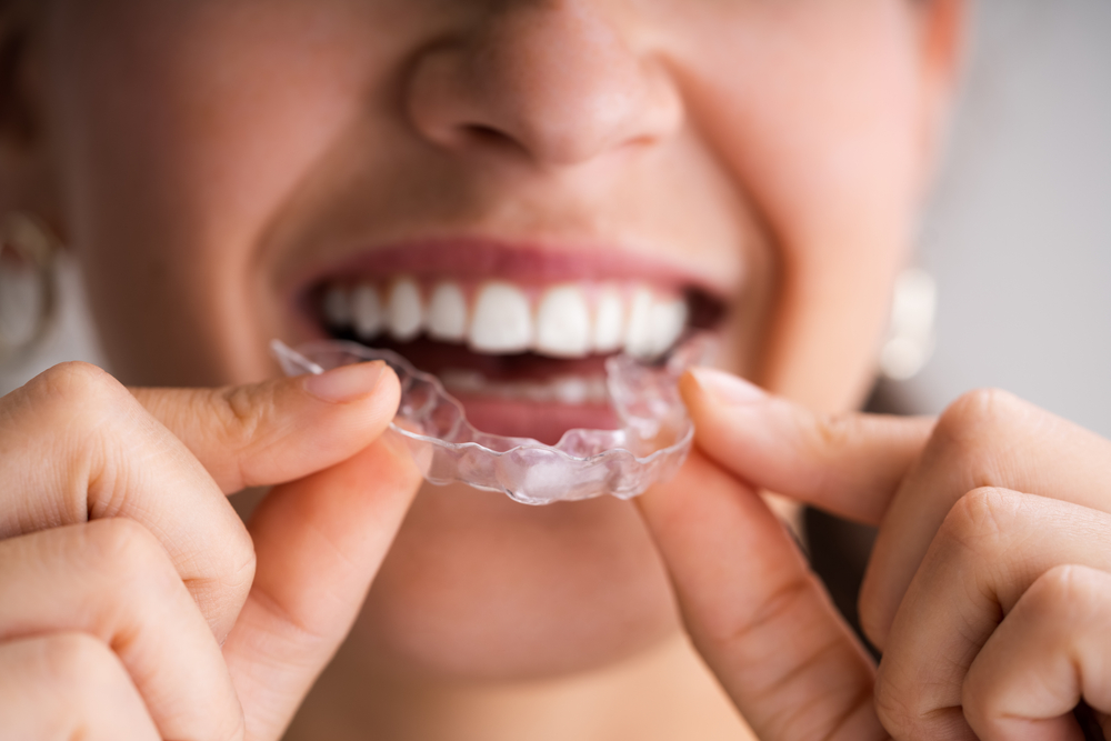 A smiling woman while holding a clear dental aligner near their teeth