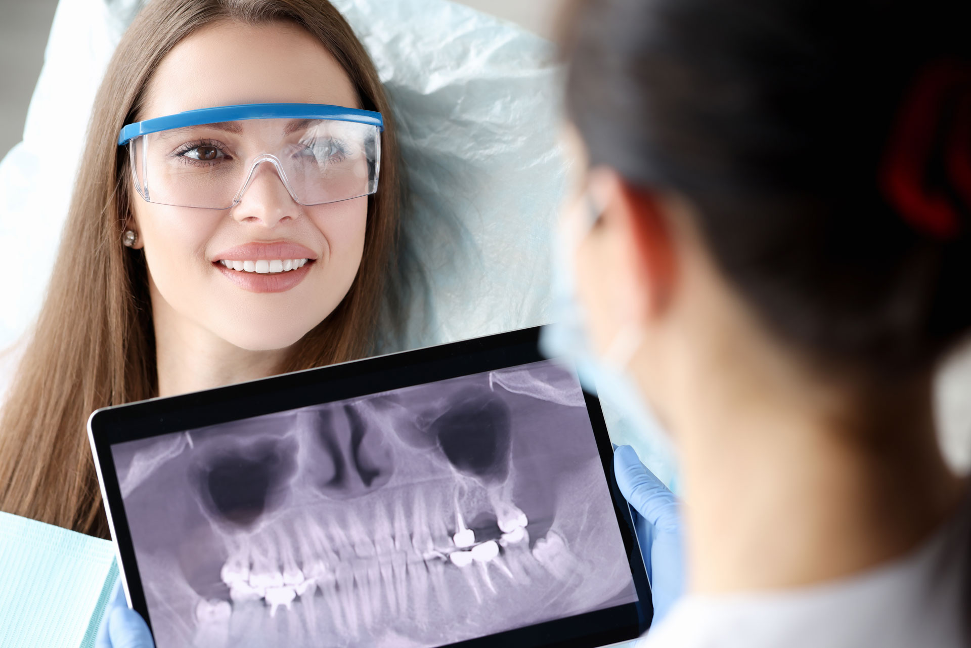 A patient wearing safety glasses looks at a dental X-ray on a tablet held by a dentist, showing a detailed image of teeth and sinuses