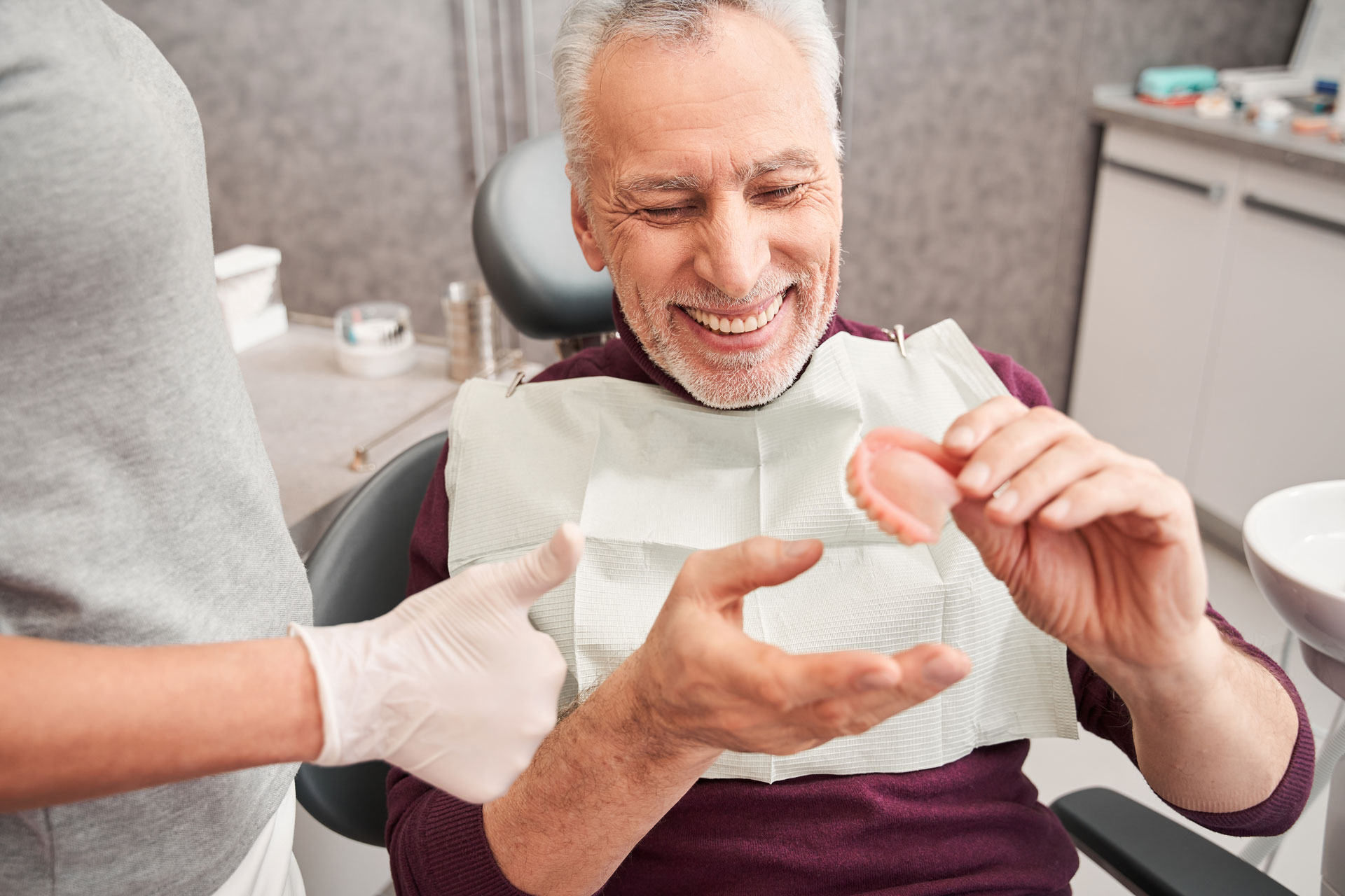 A patient with a smile sits in a dental chair holding dentures while a dental professional, wearing gloves, assists in a clinic setting