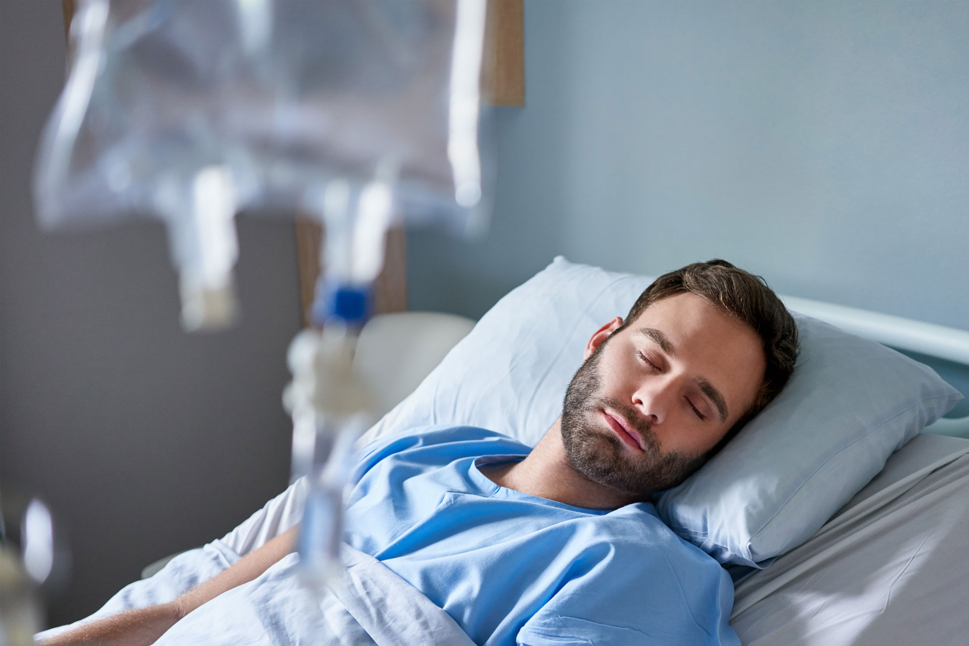 A man in a hospital bed resting with his eyes closed, while an IV drip is visible in the foreground