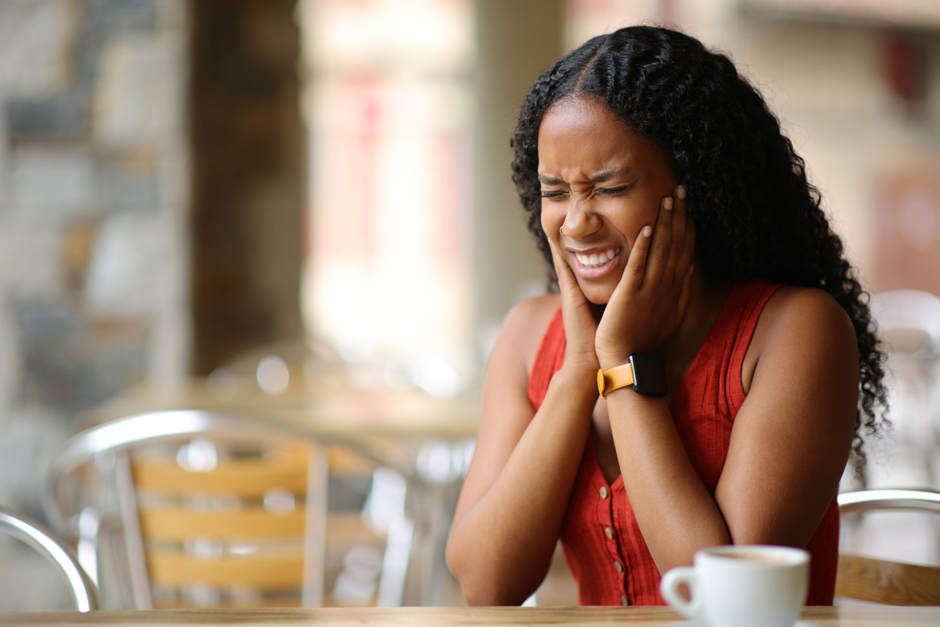 A woman in a red top sits at a table, holding her jaw in pain, suggesting symptoms of TMJ disorder