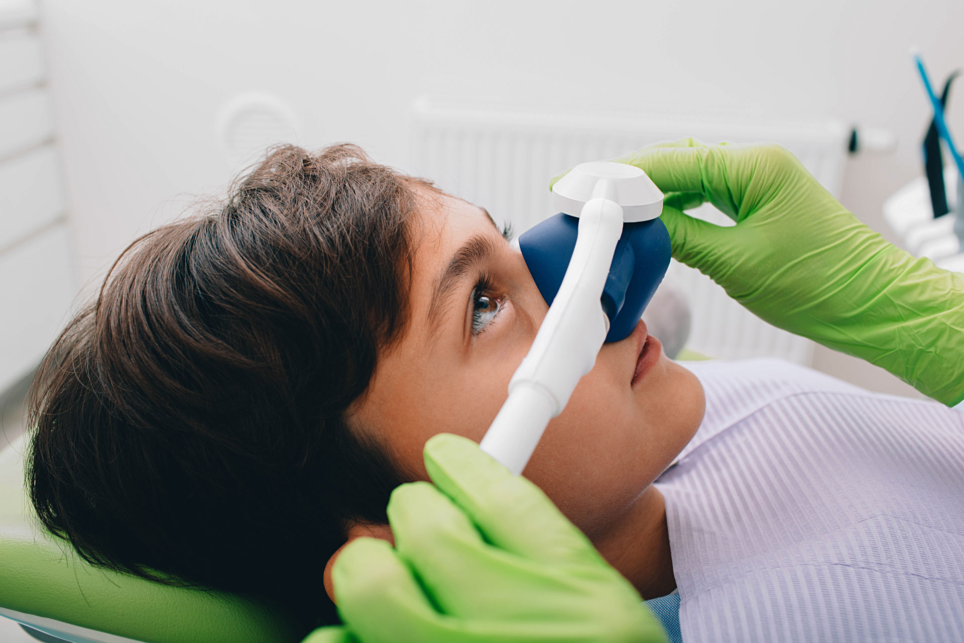 A young patient in a dental chair receives nitrous oxide through a nasal mask, while a dental adjusts the equipment