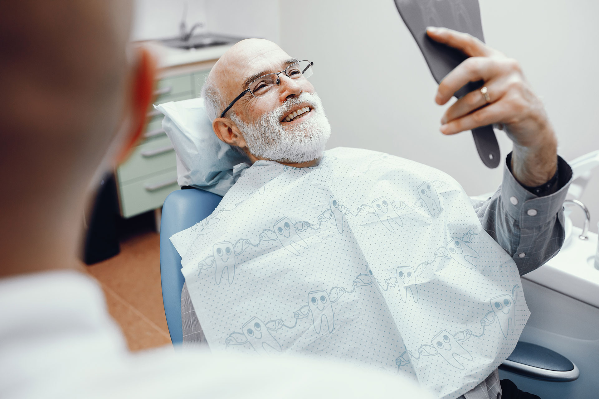 A patient smiles in a dental chair, holding a mirror and examining his teeth after reconstructive dentistry