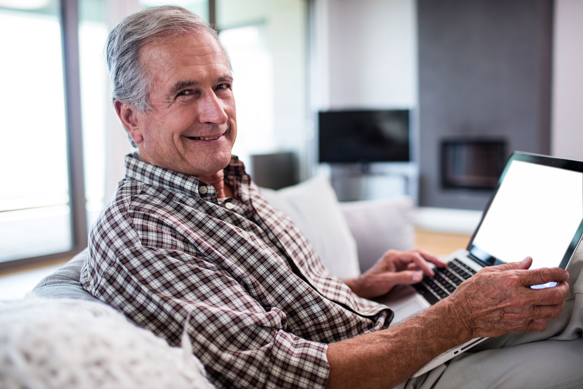 An older man sitting on a couch, smiling and using a laptop, likely completing patient forms for a dental appointment