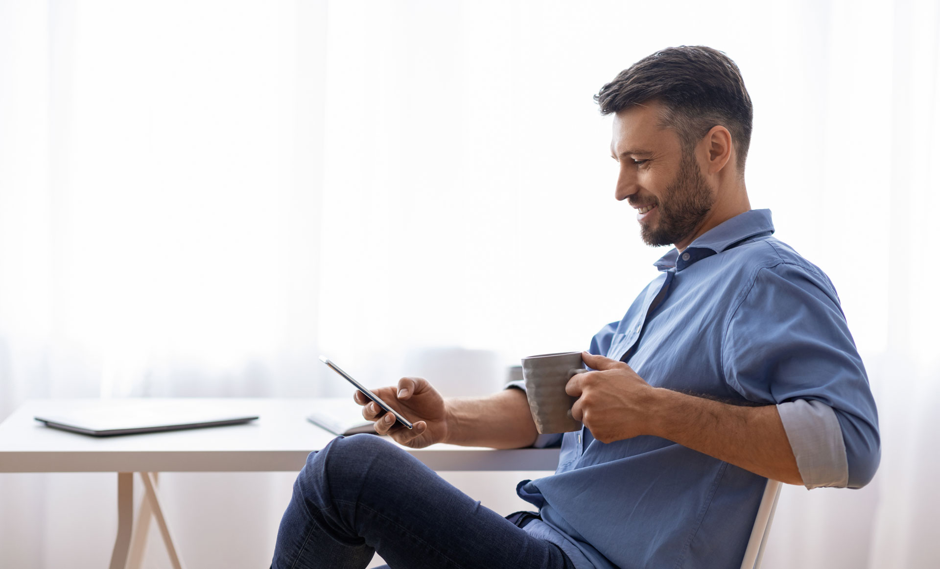 A smiling man in a blue shirt sits holding a smartphone and coffee mug, likely reviewing dental appointment details